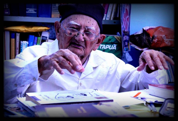 Imagem do Mosenhor Ágio Augusto Moreira sentado gesticulando com as mãos na frente de alguns livros 